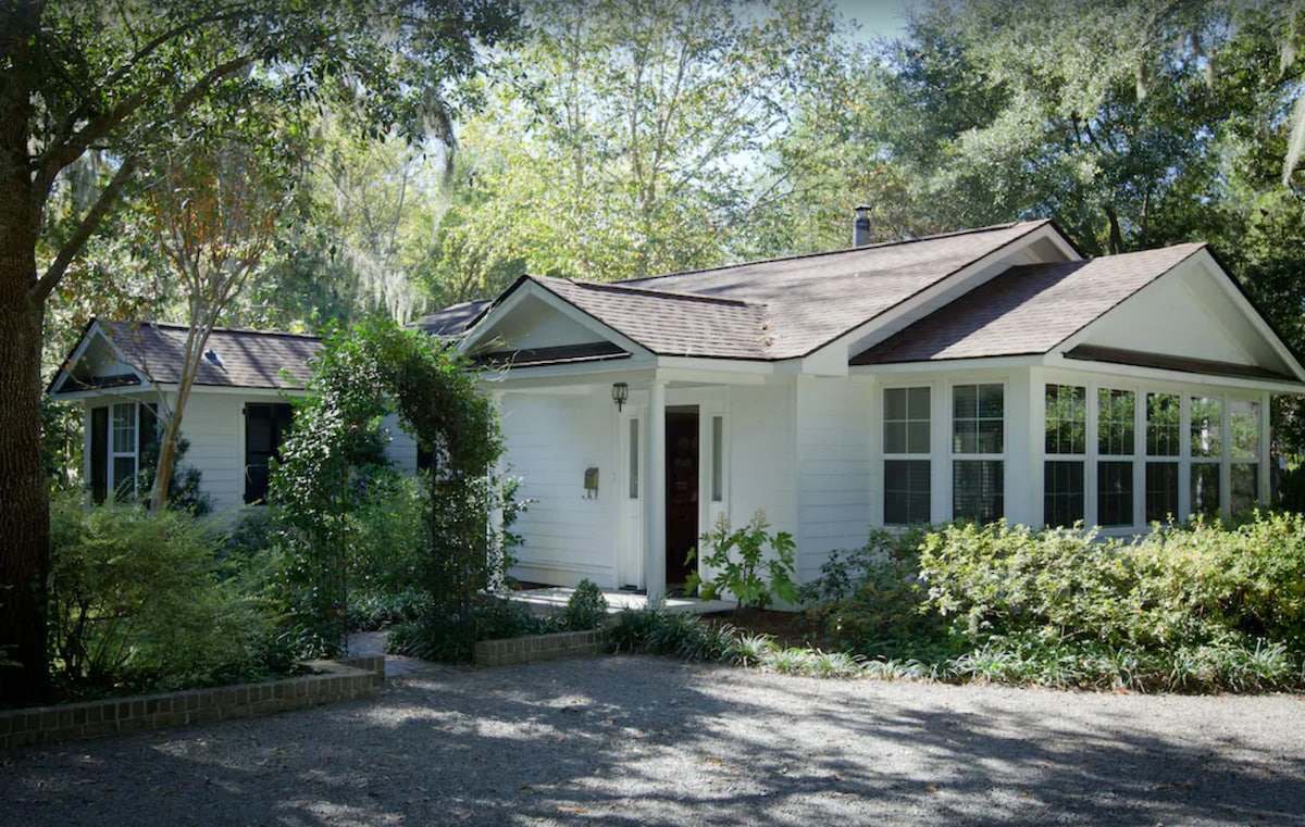 The exterior of the bungalow is showcased, featuring a charming white facade and a welcoming entrance. Lush greenery surrounds the home, with large windows allowing natural light to fill the interior. A pebbled pathway leads to the front door, enhancing the inviting atmosphere.