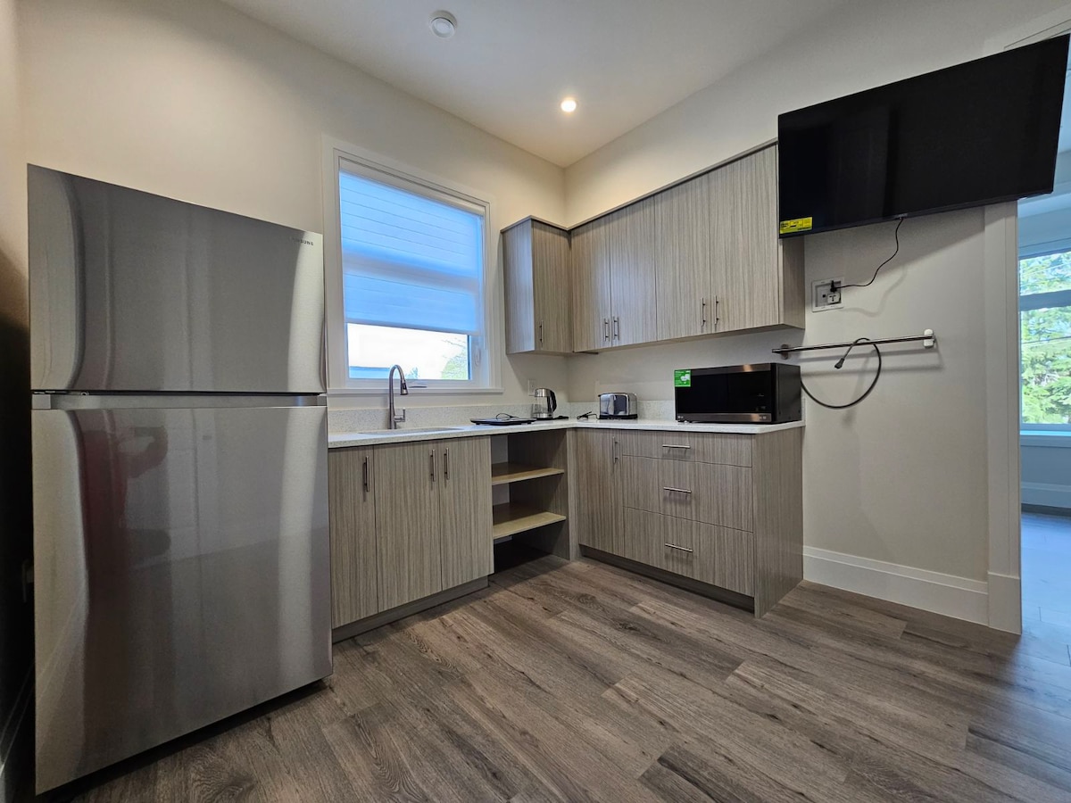 A modern kitchenette features stainless steel appliances, including a refrigerator and microwave. Wooden cabinetry provides ample storage space. Natural light streams through a sizable window, illuminating the countertop area. A flat-screen TV is mounted on the wall, enhancing functionality within the space.
