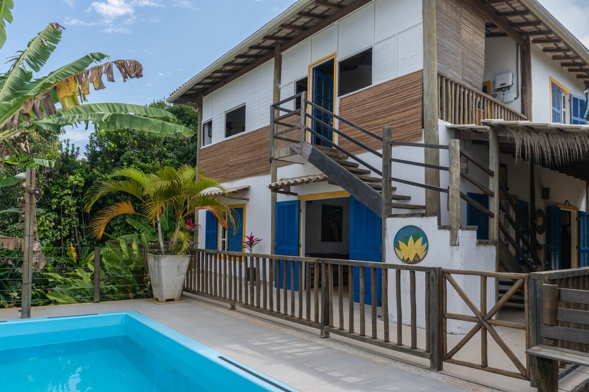 The exterior of the two-story house is displayed, featuring wooden accents and bright blue doors. A staircase leads to the upper level, while a small pool is positioned in the foreground. Lush greenery surrounds the area, enhancing the natural setting.