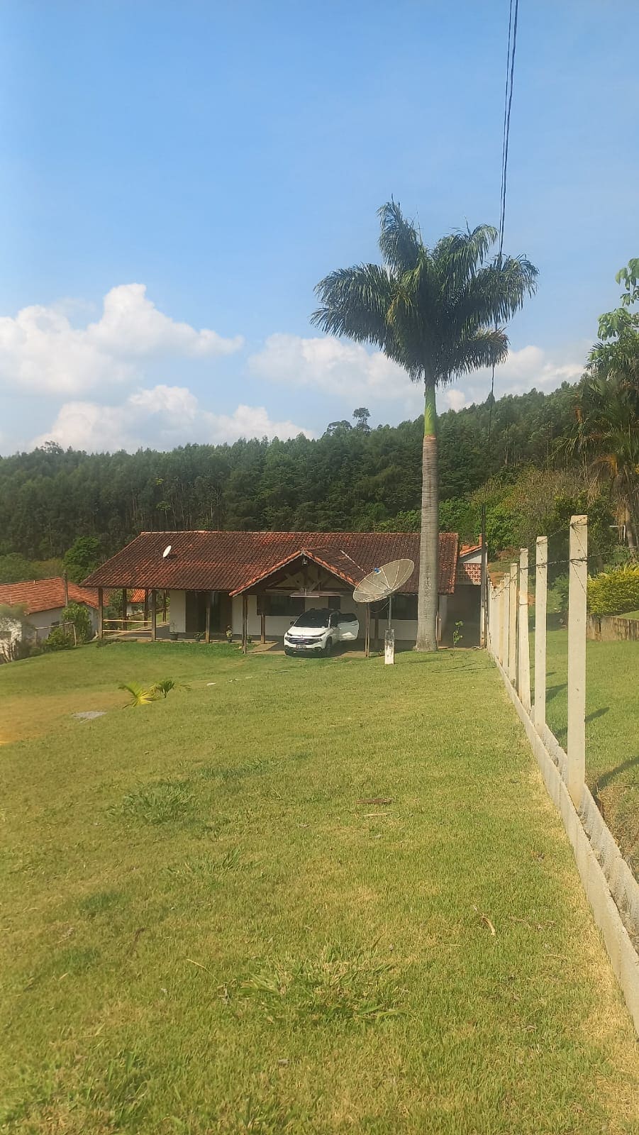 A charming single-story house is framed by a large palm tree and set against a backdrop of lush green hills. The property features a well-manicured lawn, and a parked car is visible in front of the home. The sky is partly cloudy, enhancing the serene atmosphere.