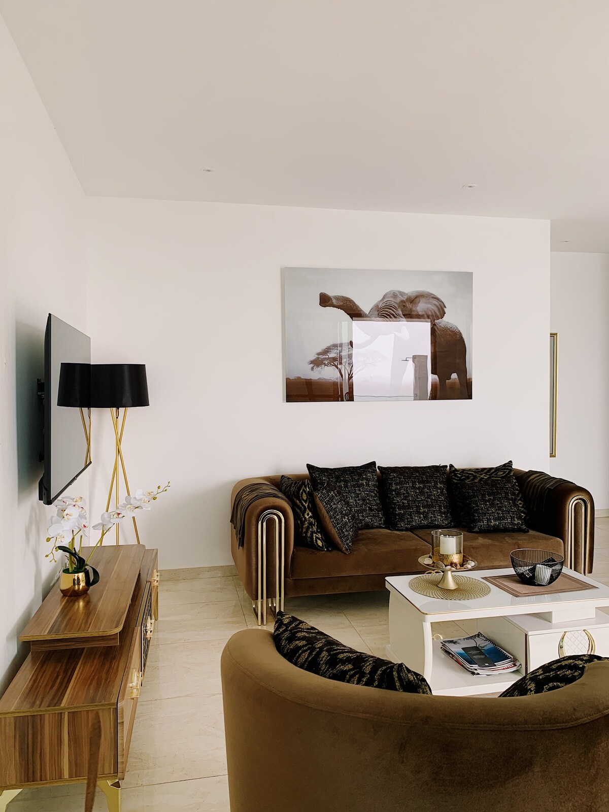 A stylish living area is featured with a plush brown sofa adorned with dark accent pillows. A sleek coffee table sits at the center, complemented by a decorative bowl. Artwork on the wall adds a contemporary touch, and a slim lamp provides soft illumination.