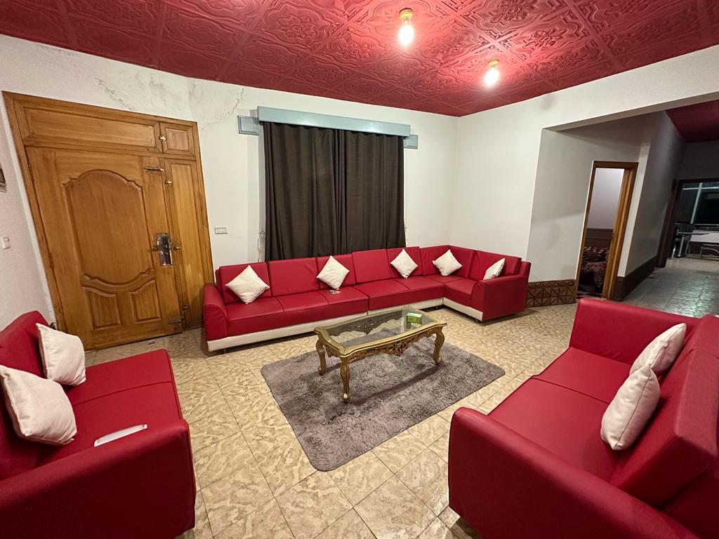 A spacious living area features a large red sectional sofa with decorative cushions, arranged around a glass coffee table. Soft lighting is provided by overhead fixtures, while a large window is covered by dark curtains, enhancing the comfortable ambiance.