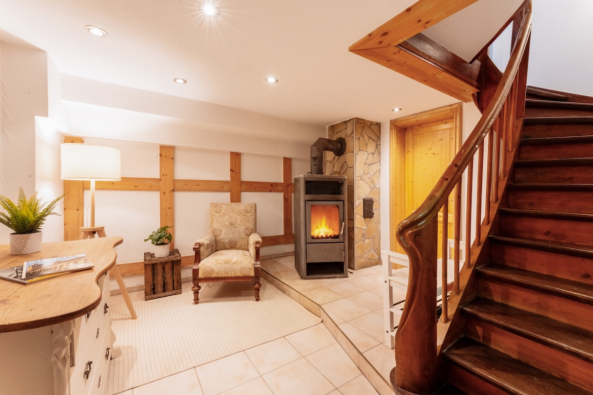 A cozy living area is highlighted by a comfortable chair positioned near a modern fireplace. The space features warm wood accents and stylish tile flooring. A staircase with wooden railings leads to the upper level. Soft lighting enhances the inviting atmosphere.