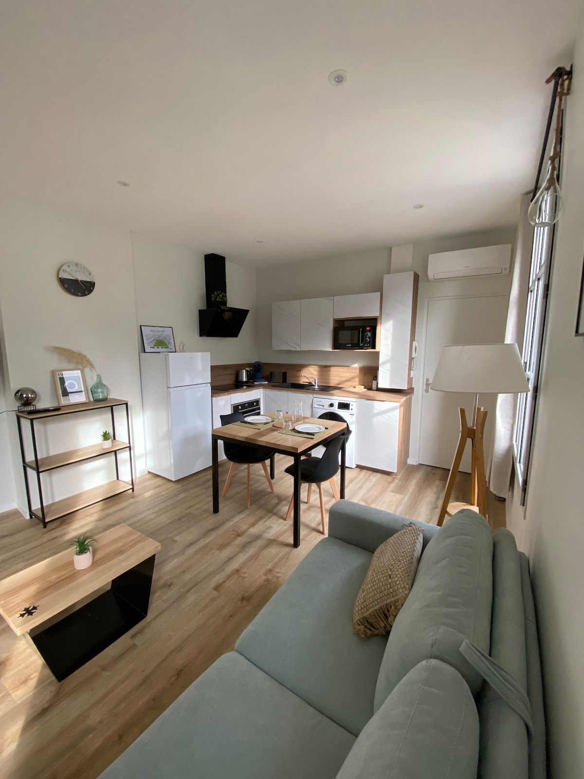 A modern open-plan living space is presented, featuring a cozy sofa and a dining table set for two. The kitchen is fully equipped with appliances, including a refrigerator, microwave, and stove. Natural light flows through the windows, highlighting the wooden floor.