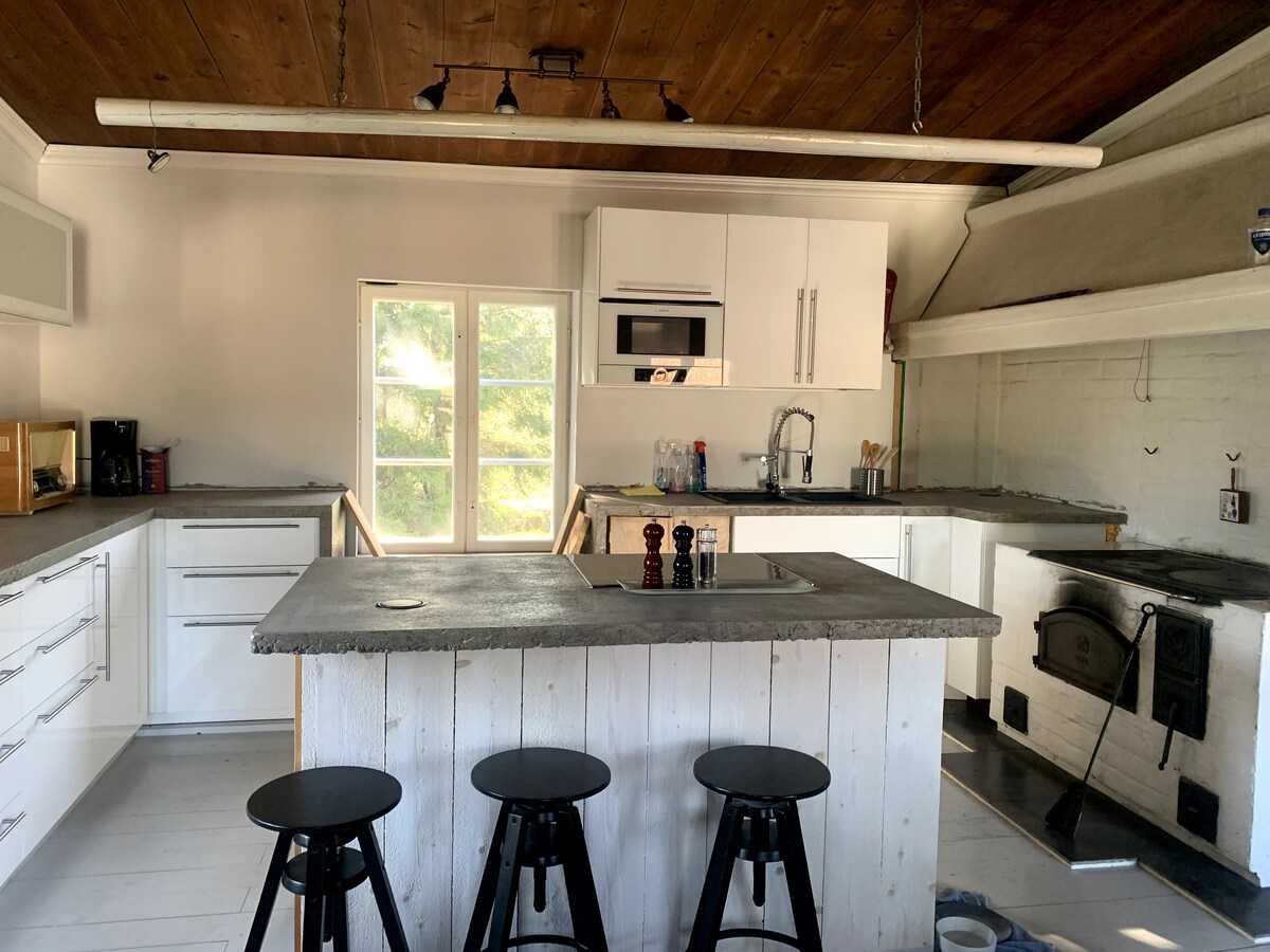A modern kitchen is showcased with white cabinetry and a concrete-topped island. Bar stools are positioned at the island. Large windows allow natural light to fill the space, highlighting the stainless-steel appliances and a vintage stove in the corner.