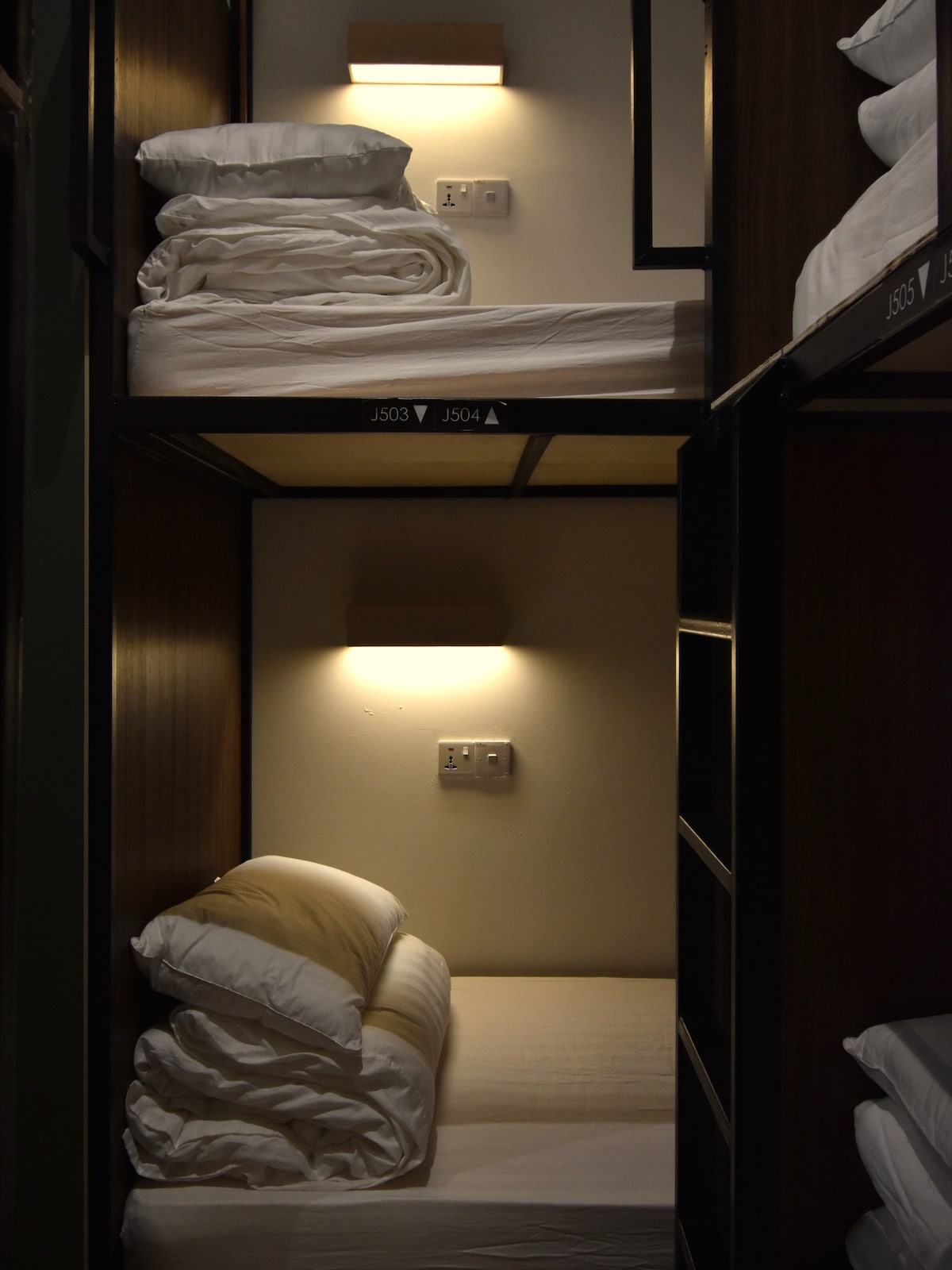 A shared dormitory room is presented with two bunk beds, each featuring neatly arranged pillows and linens. Soft lighting is provided by wall sconces above the beds, and power outlets are visible on the wall for guest convenience.