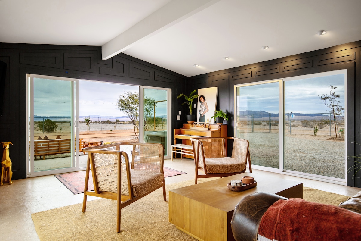 A spacious living area features large glass doors offering canyon views. Two comfortable chairs are set around a wooden coffee table, with a piano situated against a dark wall that provides a cozy contrast. Natural light fills the room, enhancing the serene atmosphere.