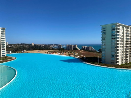 A large, inviting infinity pool stretches across the foreground, reflecting the blue sky above. In the background, modern residential buildings line the coast, with the ocean visible in the distance, enhancing the overall serene environment.