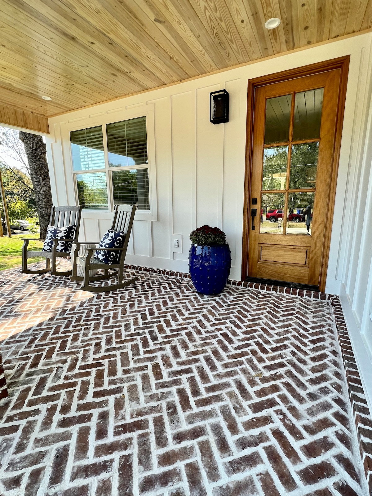A welcoming porch features a distinctive brick-patterned floor and two comfortable wooden rocking chairs. A large blue planter adds a pop of color beside the entrance, which is framed by a wooden door and a window that allows natural light to fill the space.