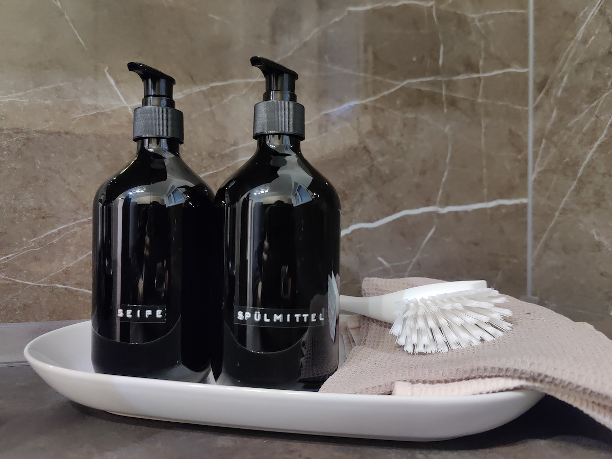 Two black bottles labeled for soap and dishwashing liquid are displayed on a sleek tray. A brush and towel are arranged neatly beside them, set against a backdrop of polished stone tiles, creating a functional and organized space.