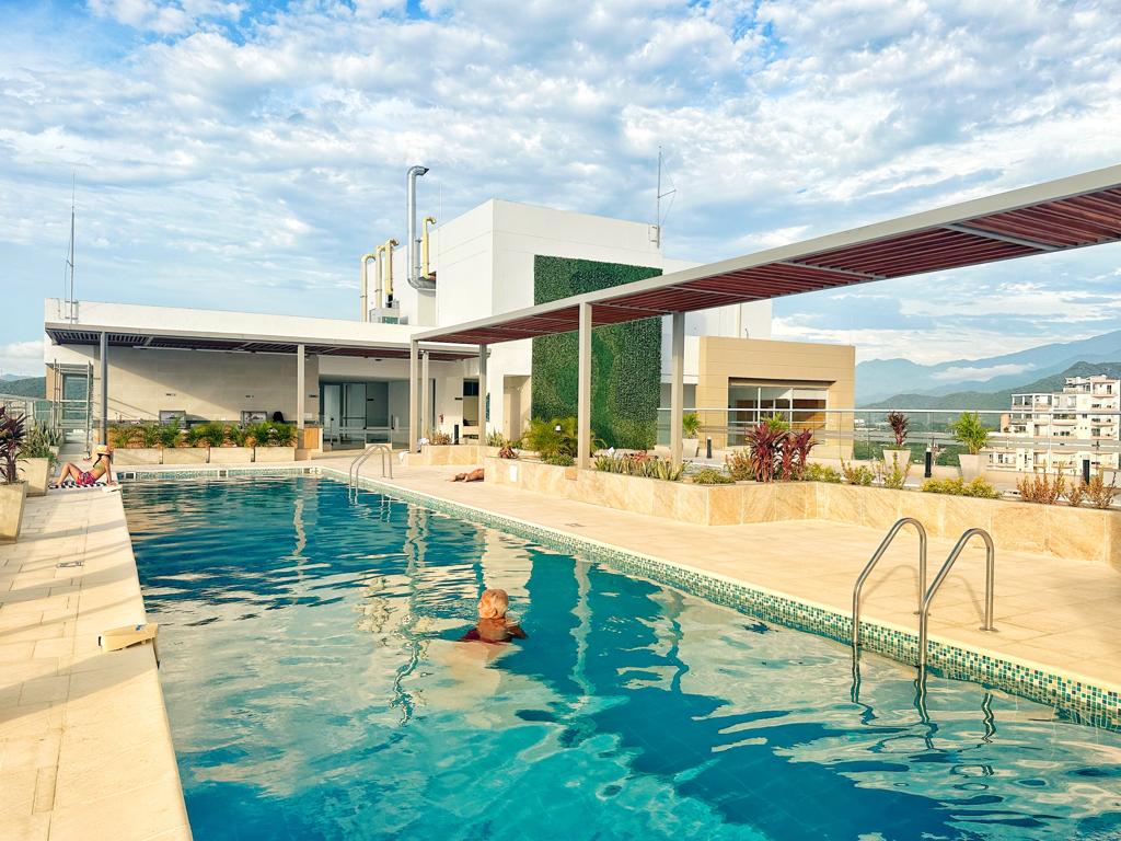 A rooftop pool is seen, featuring clear blue water and surrounded by landscaped areas. Sun loungers are positioned along the edge, while a person enjoys swimming in the inviting space. The backdrop includes modern architecture and distant mountains under a partly cloudy sky.