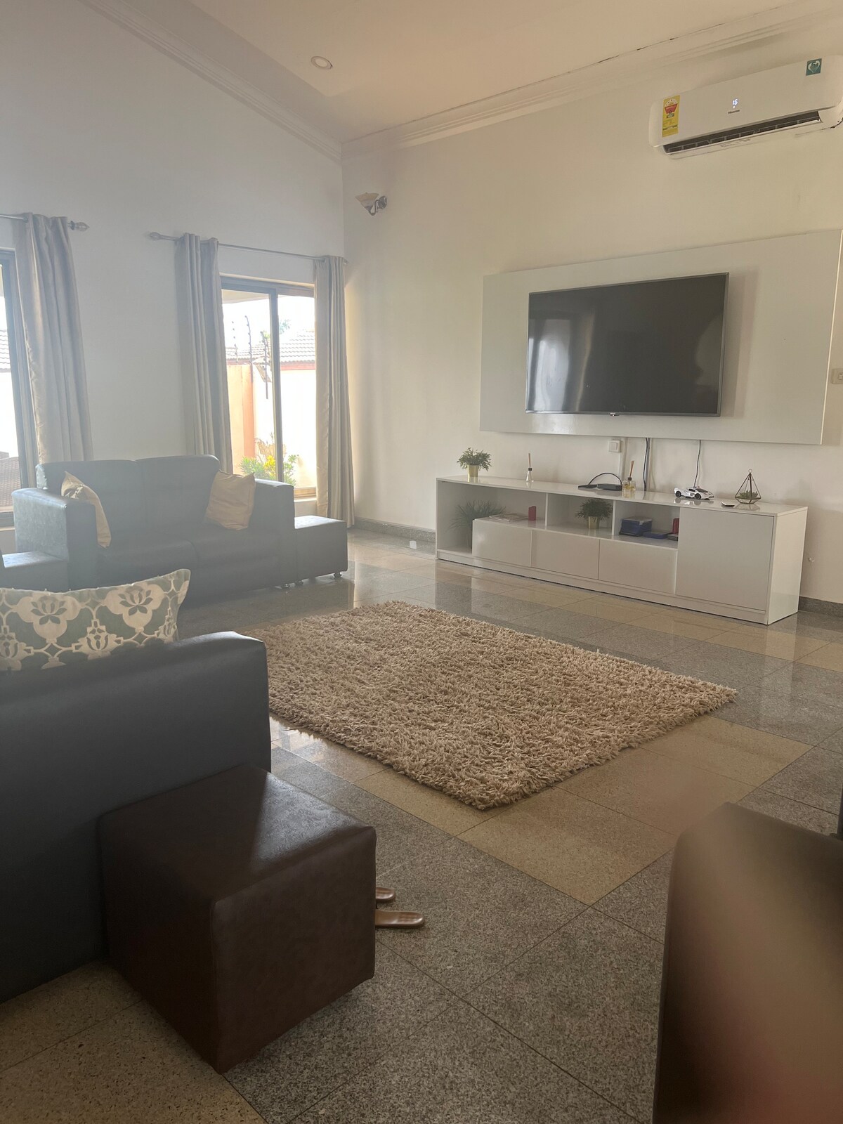 A comfortable seating area features two chairs and a sofa, complemented by a plush rug. A television is mounted on the wall above a sleek, modern console. Natural light filters in through large windows adorned with light curtains, enhancing the spacious feel.