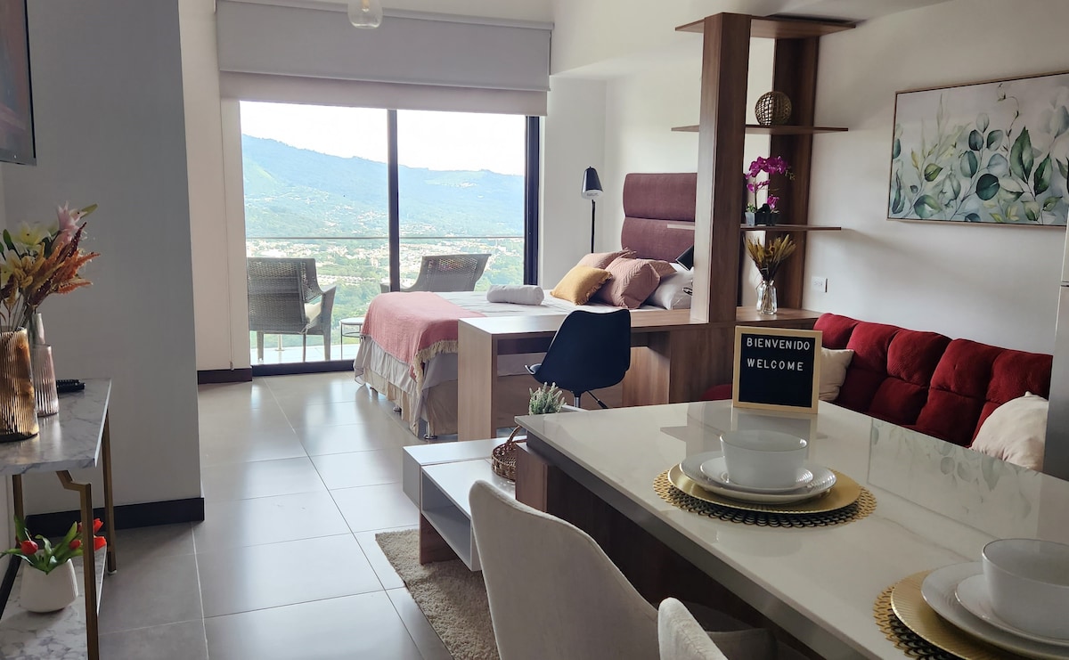 An inviting studio apartment is presented, featuring a spacious layout with a comfortable queen bed and a cozy sofa bed. A dining area with simple table settings is visible, complemented by natural light streaming through large windows overlooking the valley.