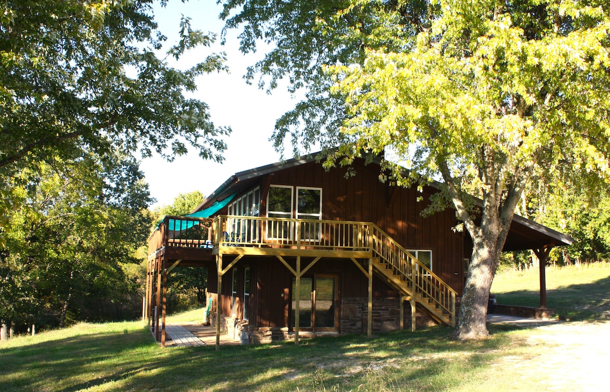 A spacious cabin is set amidst lush trees, featuring a wooden deck with a shaded area above the entrance. Stairs lead up to the deck, showcasing a large, inviting outdoor space, ideal for enjoying the expansive natural surroundings.