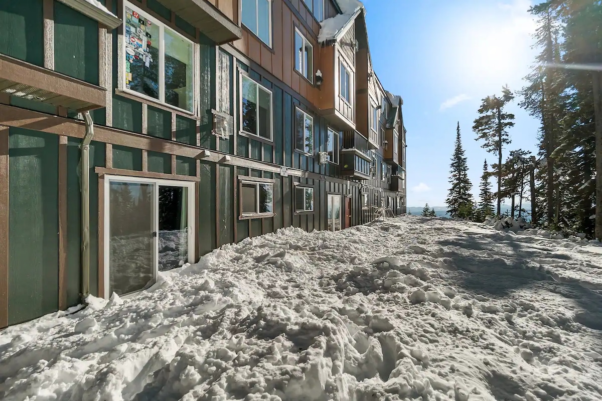 The exterior of the condo is surrounded by a thick layer of snow, creating an inviting winter atmosphere. Sunlight reflects off the snow, highlighting the building's structural design. Tall evergreen trees can be seen in the background, providing a glimpse of the picturesque alpine landscape.