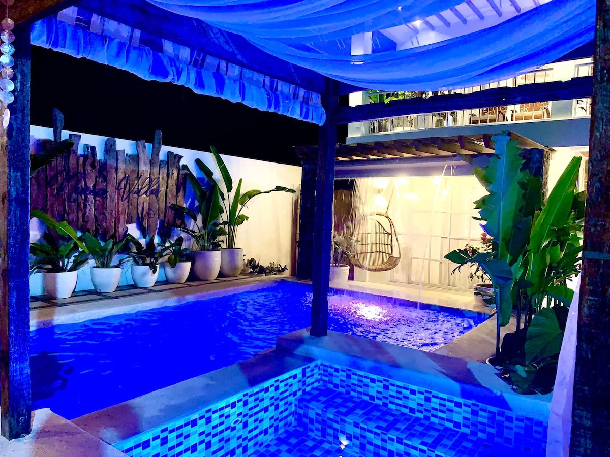 The vibrant blue pool area is surrounded by lush greenery and decorative planters. A charming jacuzzi is positioned next to the pool, under a canopy that adds shade. Soft lighting highlights the space, creating a relaxing ambience for guests to enjoy.