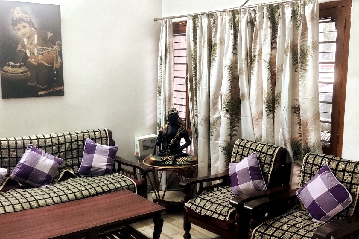 A seating area is depicted with traditional wooden chairs and a matching sofa, adorned with purple and white patterned cushions. A small round table sits in the center, while a serene statue is positioned nearby. Elegant curtains drape over the window, enhancing the cozy ambiance.