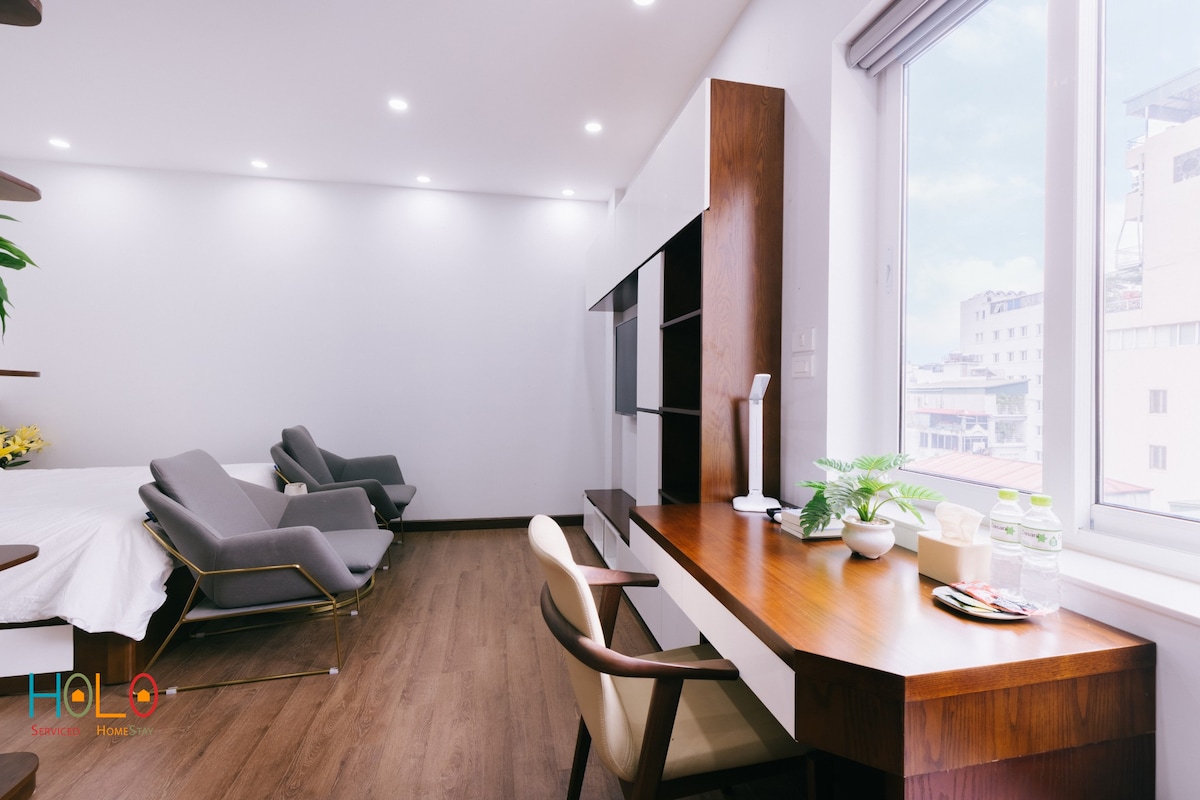 A modern apartment interior showcases a cozy sleeping area with a spacious bed and sleek gray chairs positioned near the window. A wooden desk is arranged with a plant and bottled water. Natural light filters in through the window, enhancing the room's clarity.