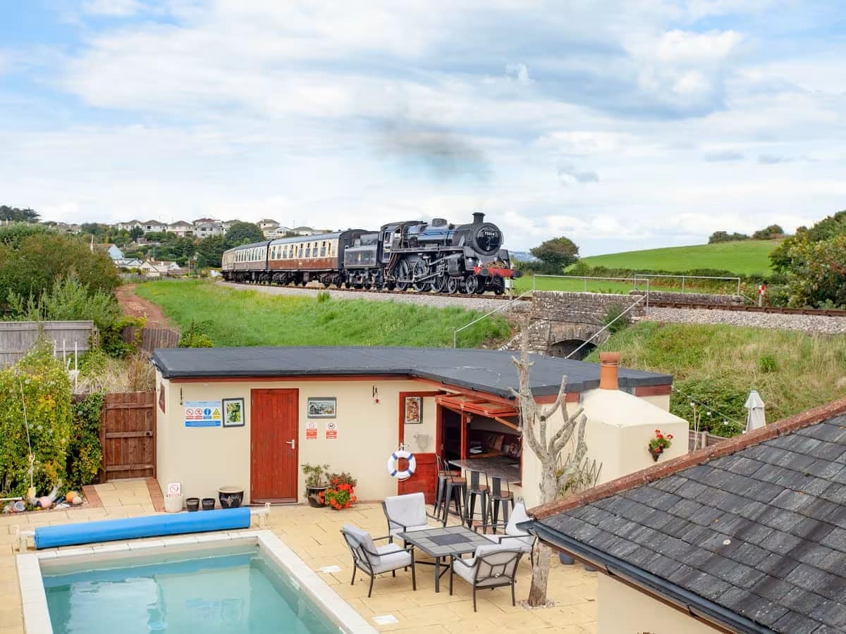 An outdoor area features a private pool surrounded by a patio and equipped with outdoor furniture. A bar area is visible to the side. In the background, a steam train travels along the railway line, flanked by green fields and distant residential buildings.