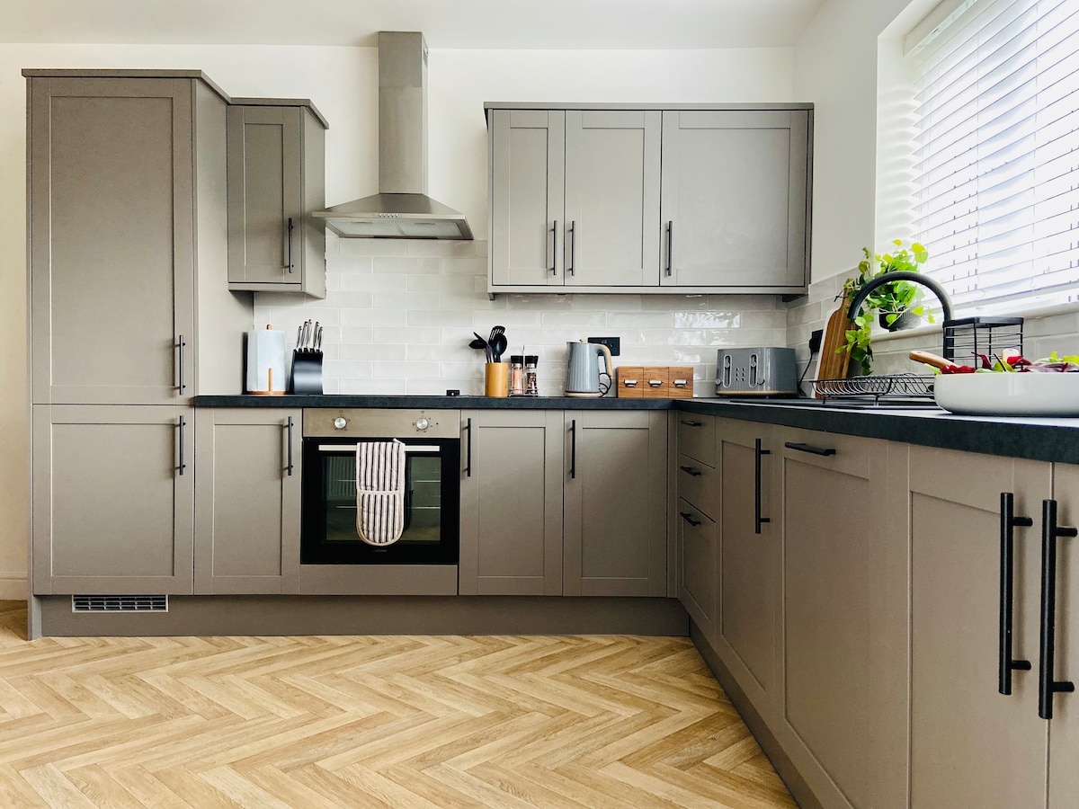 The modern kitchen features gray cabinetry, a sleek stove, and a mix of appliances including a kettle and toaster. A spacious counter provides ample workspace, while a bowl of fresh fruit adds a pop of color. Natural light filters through the window, illuminating the wood-patterned flooring.