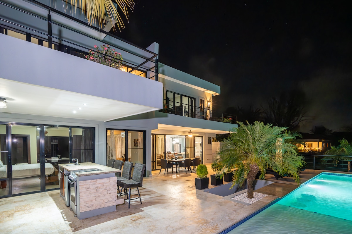 The villa's exterior is illuminated at night, displaying a spacious terrace with outdoor seating and a bar area. A private swimming pool reflects the surrounding palm trees and architecture, while large glass doors provide a glimpse into the interior living spaces.