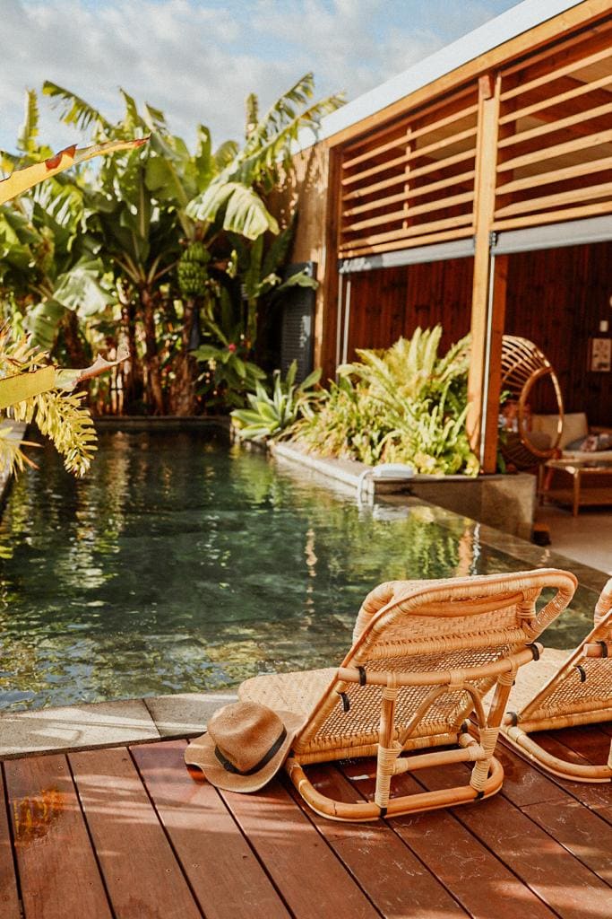 Two rattan chairs are positioned near a tranquil infinity pool surrounded by lush tropical plants. Natural wood decking stretches alongside the water, offering a serene outdoor space. Soft sunlight filters through the greenery, enhancing the relaxing atmosphere.