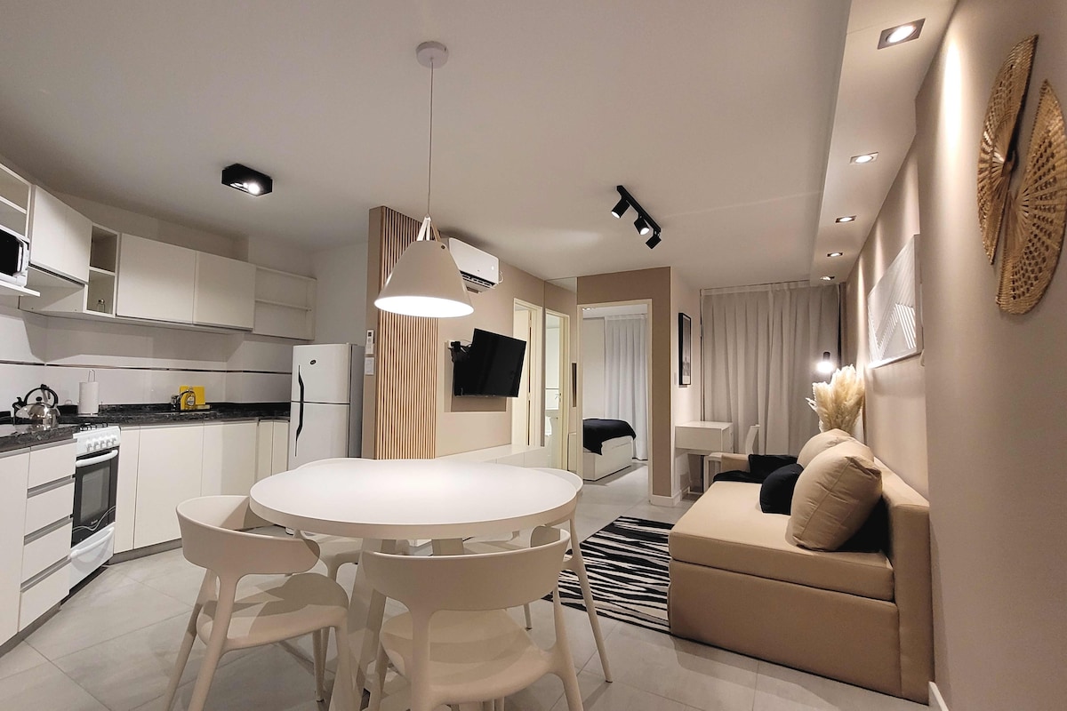 A modern living space features a circular dining table surrounded by white chairs, complemented by a beige sectional sofa. The kitchen area displays sleek, white cabinetry and stainless steel appliances. Soft lighting highlights the neutral-toned decor, creating a bright and airy atmosphere.