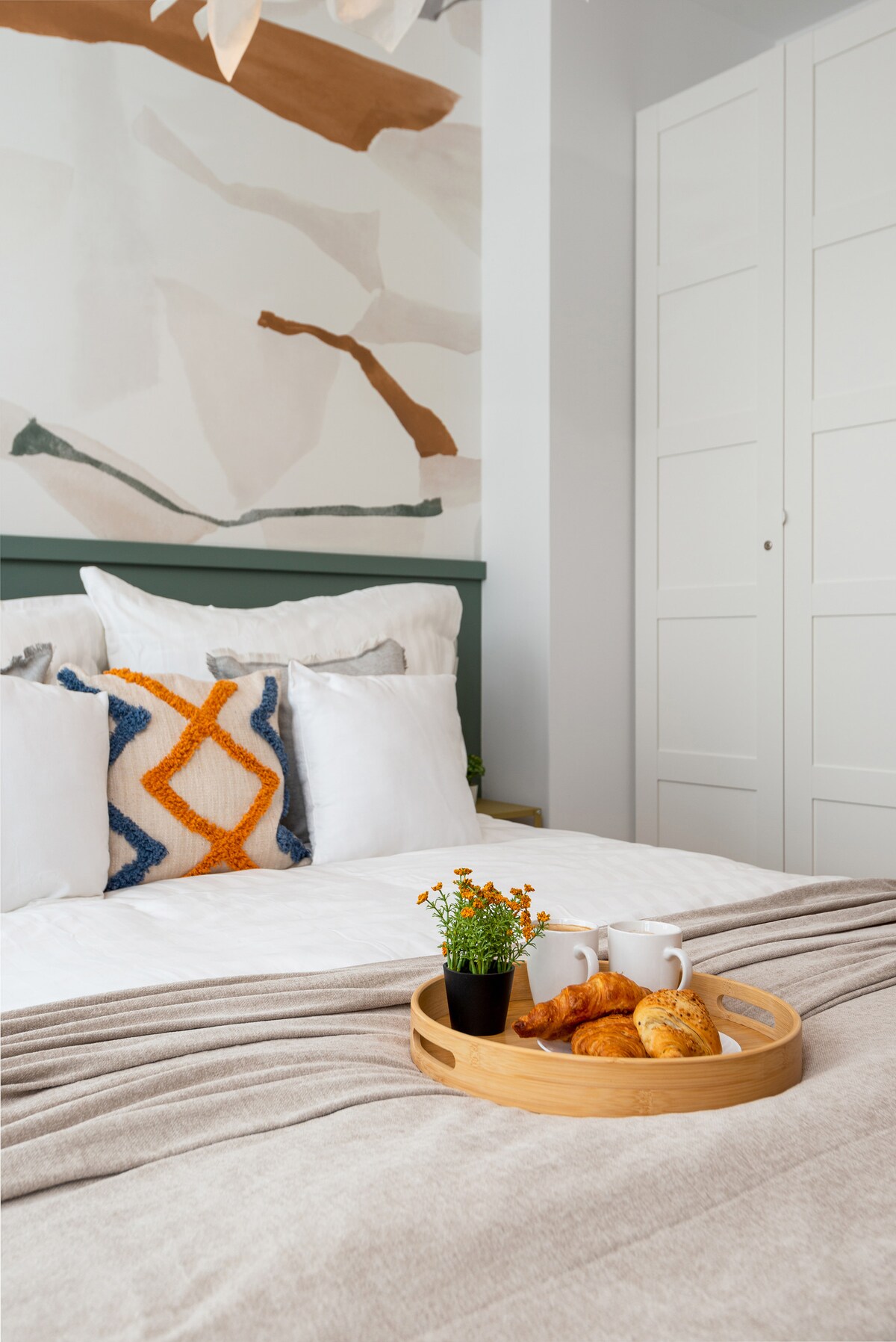 A well-appointed bedroom features a spacious bed adorned with fluffy pillows and a patterned decorative pillow. A wooden tray holds beverages and baked goods, while a small potted plant adds a touch of greenery. A stylish mural enhances the room's visual appeal.