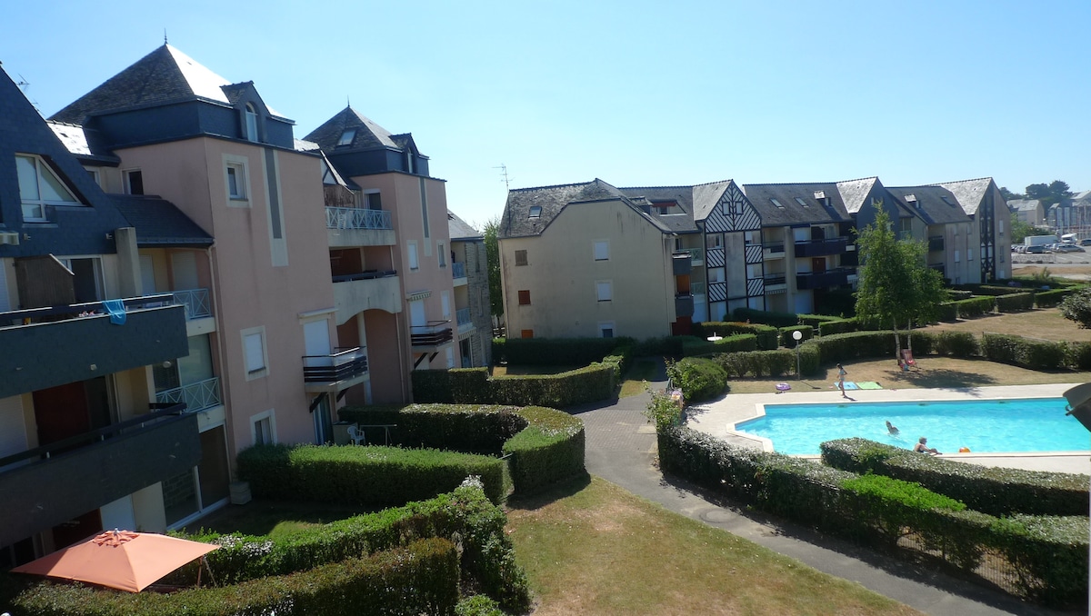 An overview of a residential complex featuring multiple buildings with varying architectural styles. Surrounding the property are neatly trimmed hedges and a swimming pool, with guests enjoying the water under clear blue skies. The image highlights a sense of community and leisure.