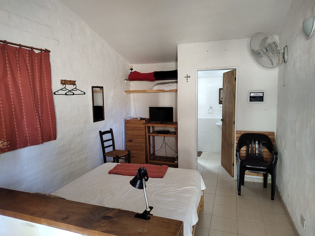 A simple room features a spacious bed covered with a white blanket. A small desk with a lamp and a dresser with a television are present. Natural light enters through a window dressed with red curtains, while a chair and a hanging storage rack complete the space.