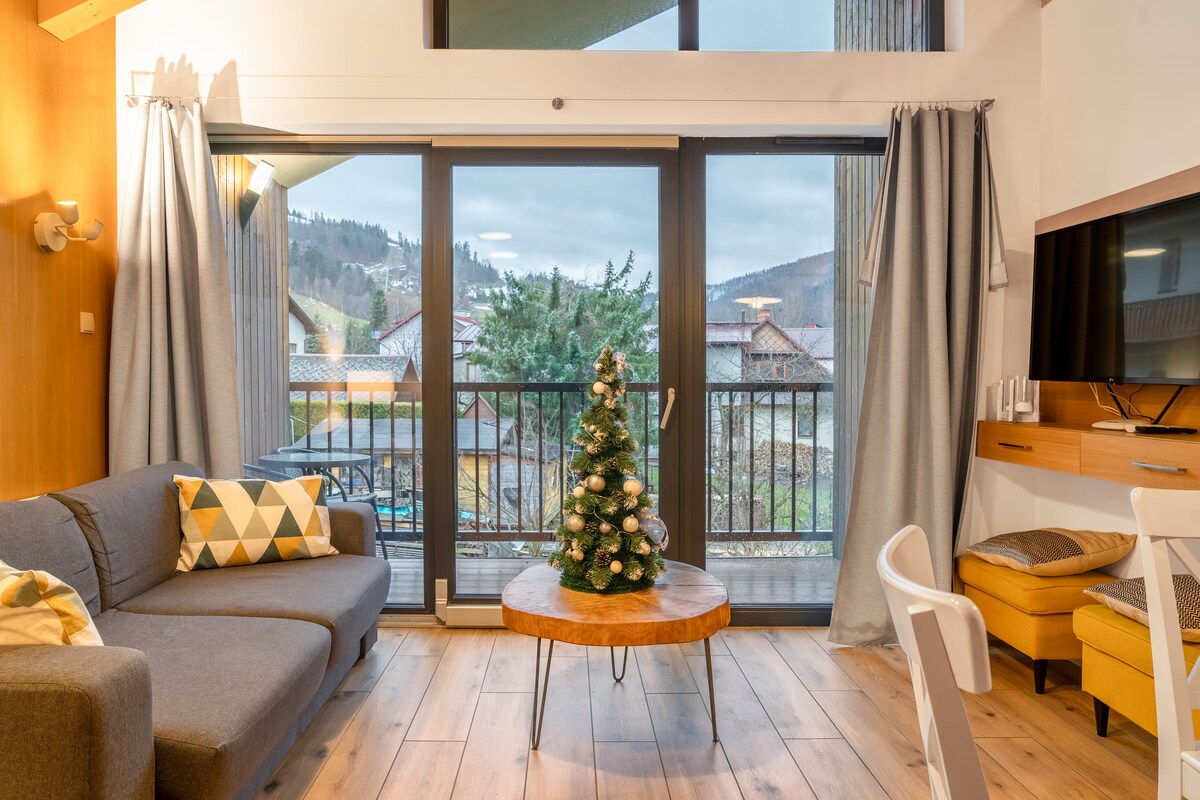 A living area features large glass doors opening to a balcony, allowing natural light to fill the space. A cozy sofa and wooden coffee table are complemented by a small decorated Christmas tree. Light-colored curtains frame the windows, enhancing the room's spacious feel.
