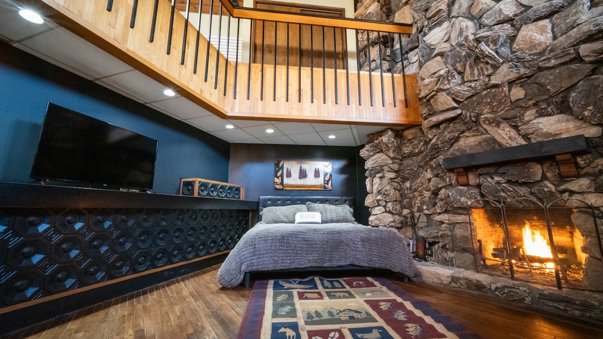 A warm sleeping area features a spacious bed positioned in front of a stone fireplace. A large television is mounted on the wall, with a patterned area rug adding texture to the warm wood floor. A wooden balcony above adds to the open feel.