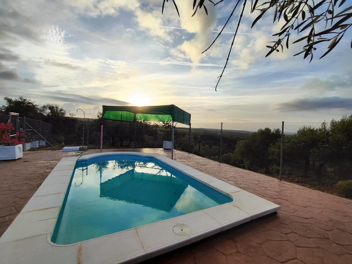 A secluded pool area is captured, featuring a rectangular pool bordered by a light-colored stone deck. A green awning provides shade, while the sunset casts a warm glow over the landscape, showcasing rolling hills and greenery in the background.