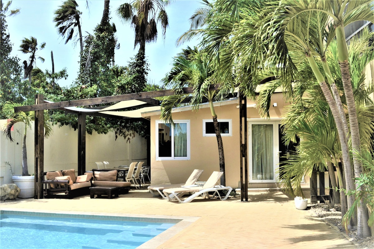 The outdoor area features a large pool surrounded by palm trees. Comfortable lounge chairs and two outdoor seating areas are positioned under a pergola, providing a shaded space for relaxation. The bright exterior of the apartment complements the tropical setting.