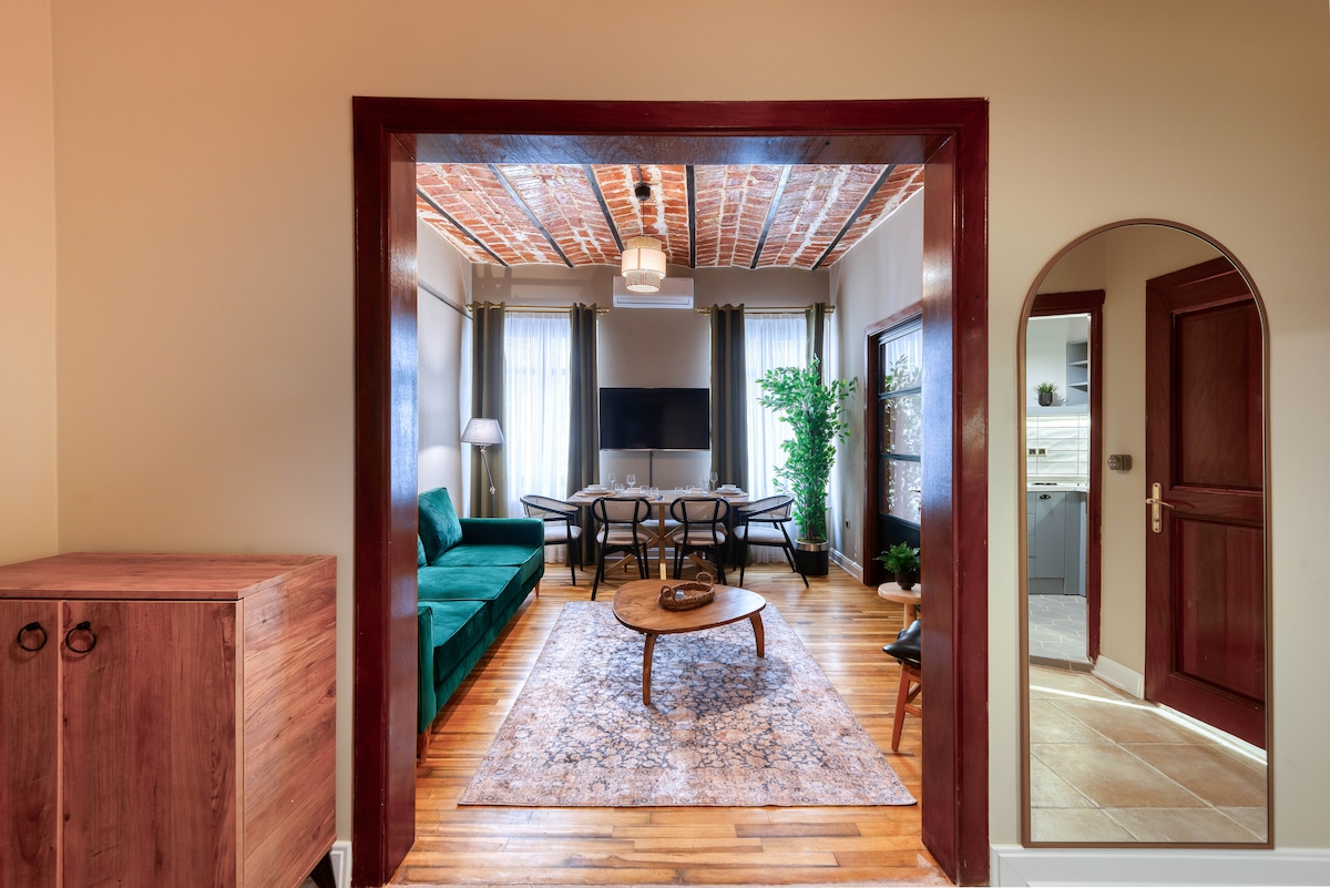 A spacious living area features a green sofa and a large dining table. Natural light filters through the tall windows, and a decorative area rug enhances the wood floor. A subtle plant adds a touch of greenery, with a wooden cabinet and an arched mirror framing the entrance.