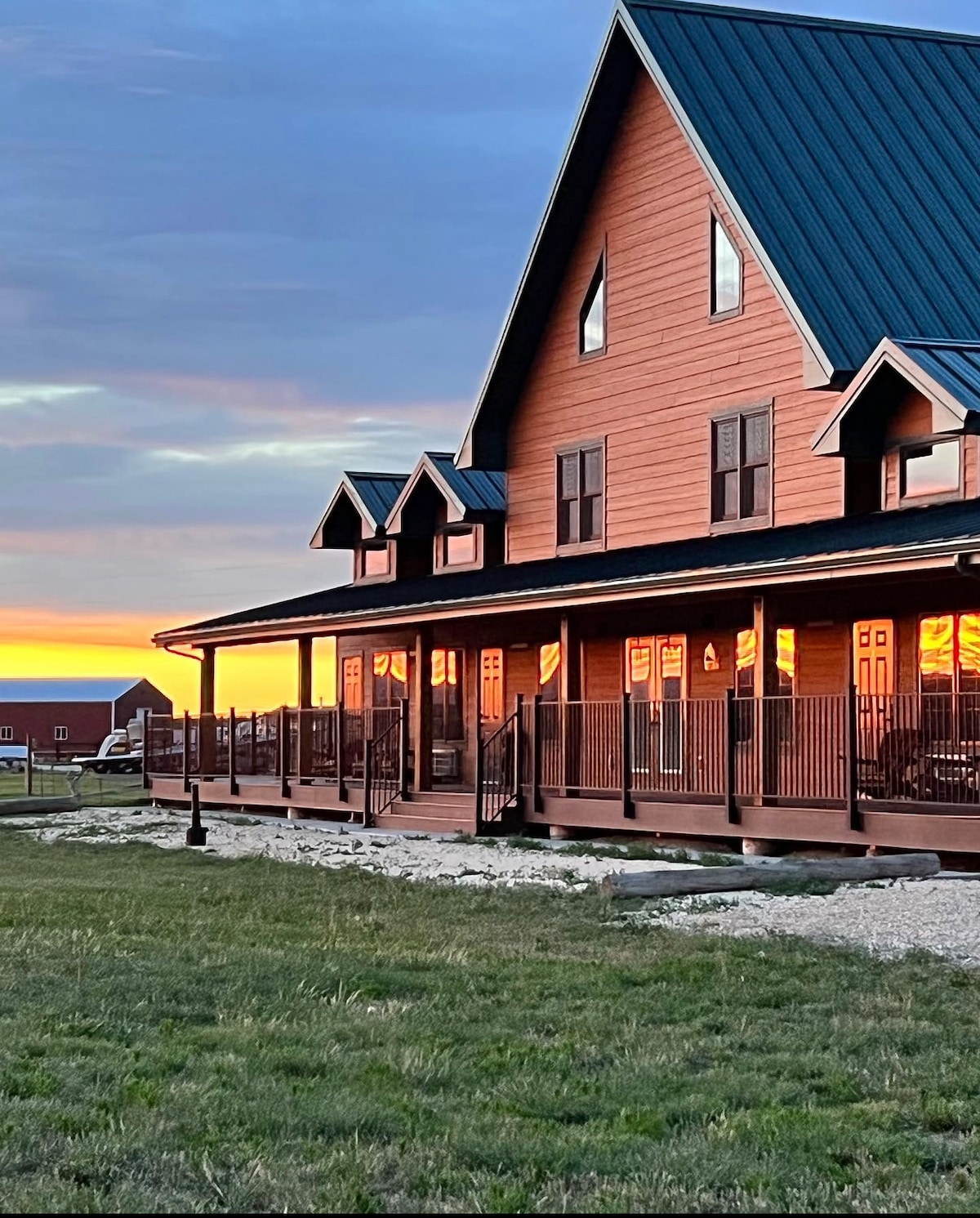 A spacious two-story house is presented with a wooden exterior and a steeply pitched roof. Large windows reflect the warm colors of the sunset, while a wraparound porch offers an inviting outdoor space. The grassy area surrounds the home, creating a serene environment.