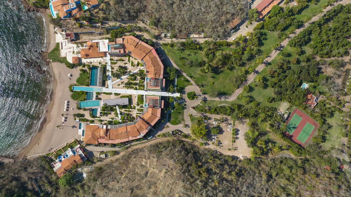 An aerial view captures the layout of El Careyes Club and Residences, showcasing a cluster of buildings surrounded by lush greenery and pathways. The beachfront is visible to the left, with pools and amenities integrated into the landscape, complemented by a tennis court nearby.