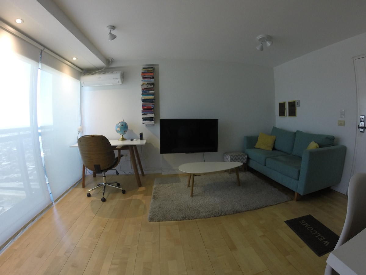 A modern living area features a light blue sofa and a white coffee table on a soft rug. A desk with a globe sits opposite a wall-mounted television. Tall windows allow natural light to fill the space, highlighting the wooden flooring.
