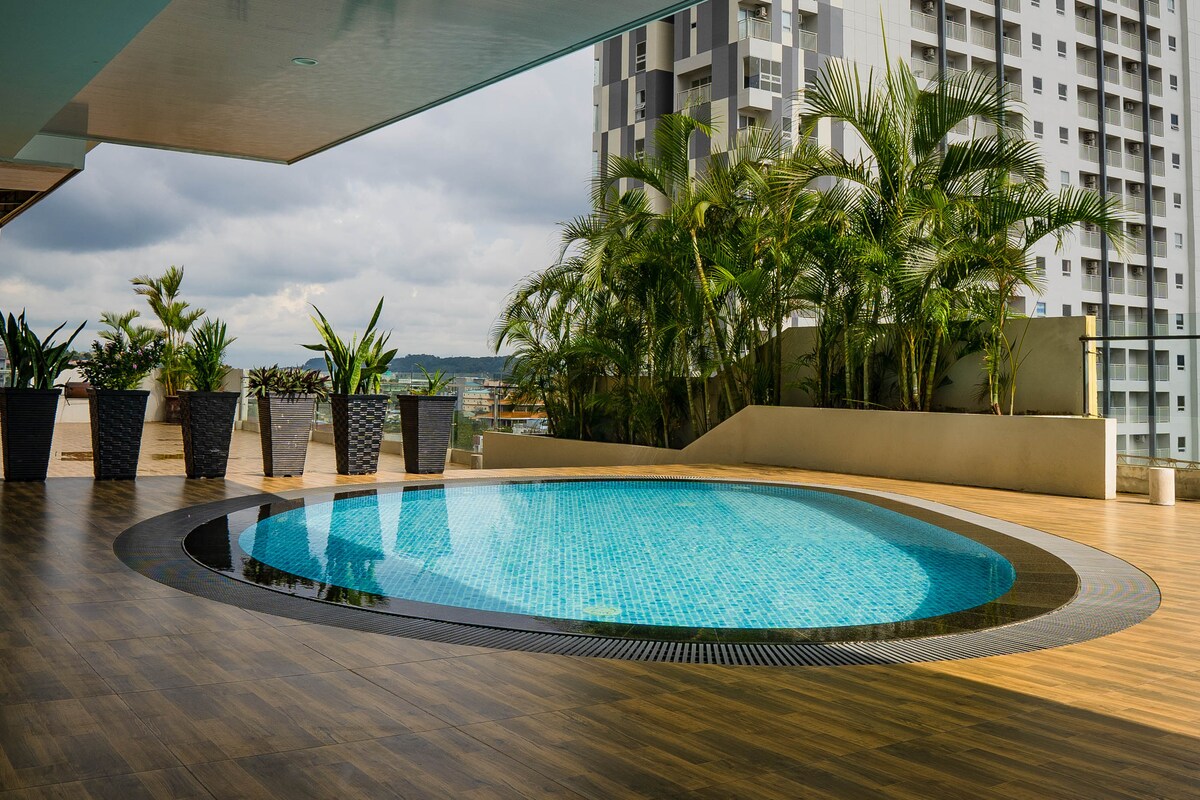 A circular swimming pool is set within a spacious outdoor area. Surrounding planters filled with lush greenery enhance the environment. Grey skies and tall buildings in the background create a serene view, with wooden decking providing a warm contrast beneath.