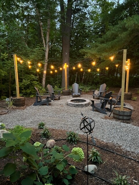 An outdoor firepit area is surrounded by gravel and soft lighting suspended from wooden posts. Several Adirondack chairs are arranged neatly around the firepit, enhancing the inviting atmosphere. Lush greenery and decorative plants add a natural touch to the secluded space.