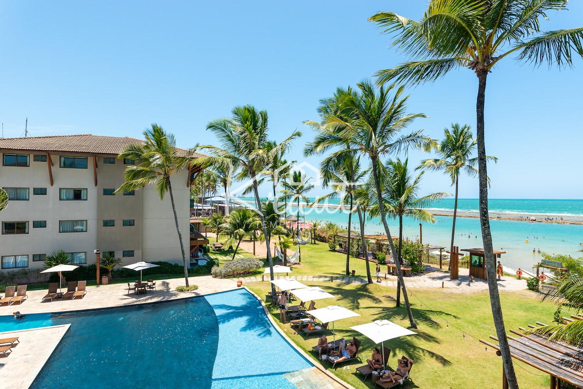 The image displays a well-maintained pool area bordered by lush palm trees, with loungers arranged under shaded umbrellas. A beachfront view is seen in the background, showcasing the clear blue ocean and sandy coastline. The vibrant sky enhances the inviting atmosphere of the location.