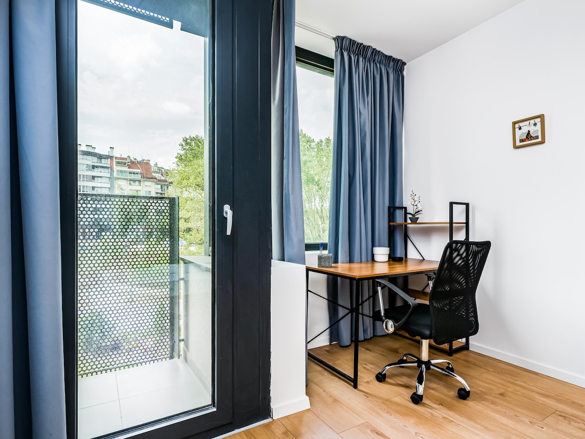 A work desk with a black chair is positioned near a large window, allowing natural light to fill the space. Sheer blue curtains frame the window, which overlooks the surrounding area. A small decorative piece is displayed on the desk, enhancing the functional space.