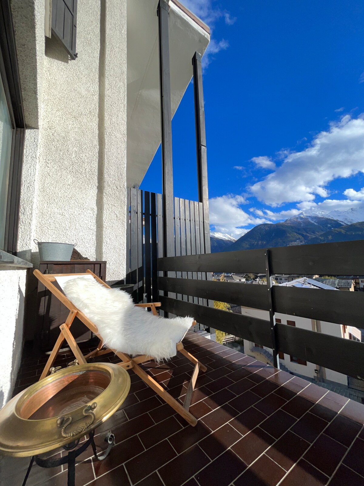 A spacious balcony features a wooden folding chair with a plush white throw, inviting relaxation. A round metal table sits beside it, and a wooden railing provides a barrier with stunning mountain views and a bright blue sky in the background.