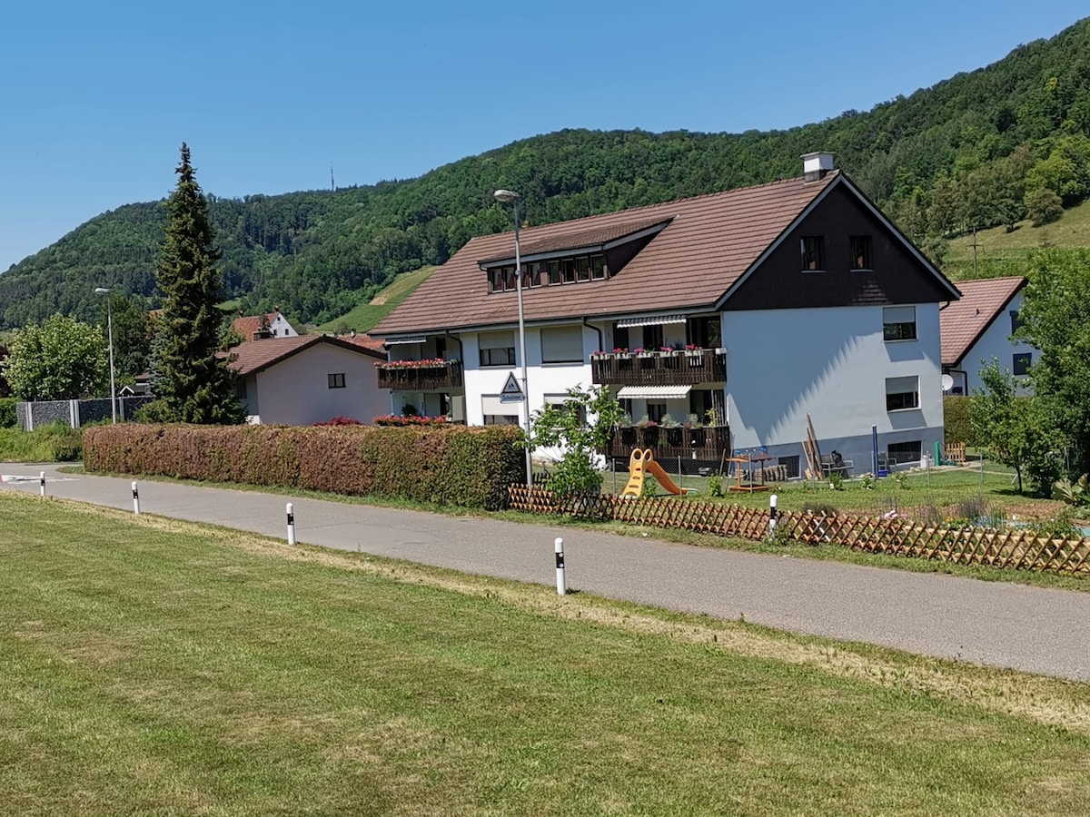 The exterior of the building showcases a modern structure nestled among greenery. A spacious lawn is seen in the foreground, with a pathway leading up to the property. Balconies decorated with plants are visible on the upper level, alongside tree-covered hills in the background.
