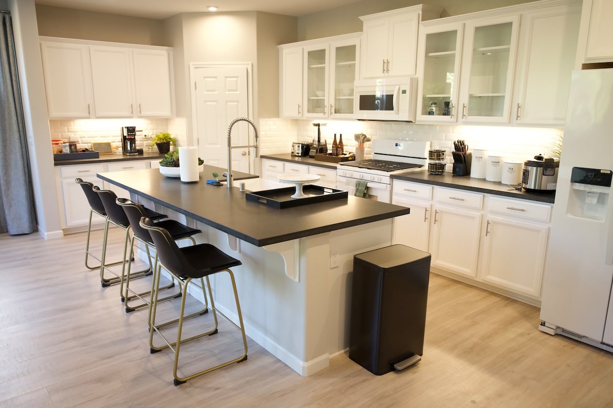 A modern kitchen features sleek white cabinetry and a dark countertop island with seating for four. Stainless steel appliances and a well-organized layout are visible, including a microwave and oven. Under-cabinet lighting enhances the workspace, while light wood flooring completes the contemporary design.