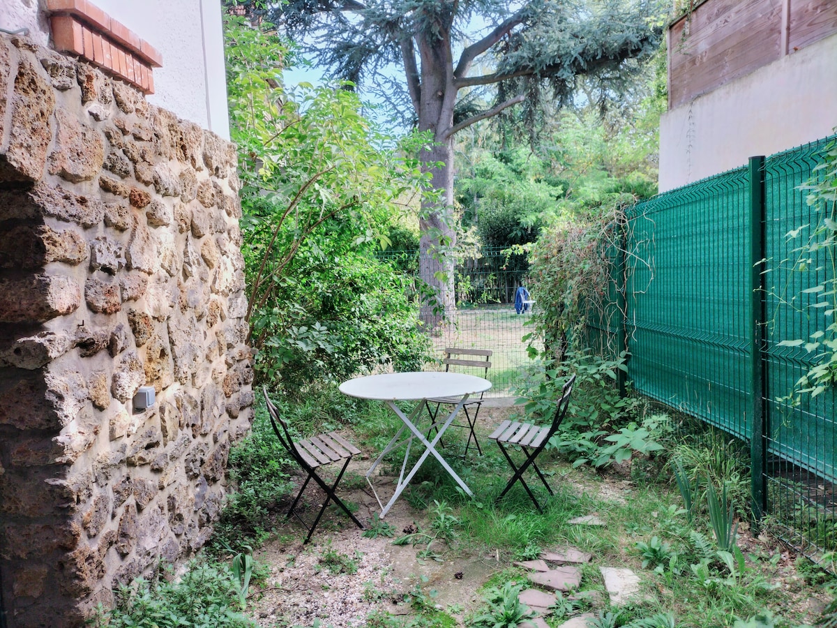 A private garden area features a round white table surrounded by two foldable chairs. Lush greenery and plants create a natural enclosure, while a stone wall provides texture. A tree stands nearby, and a fenced boundary ensures privacy in this tranquil outdoor space.