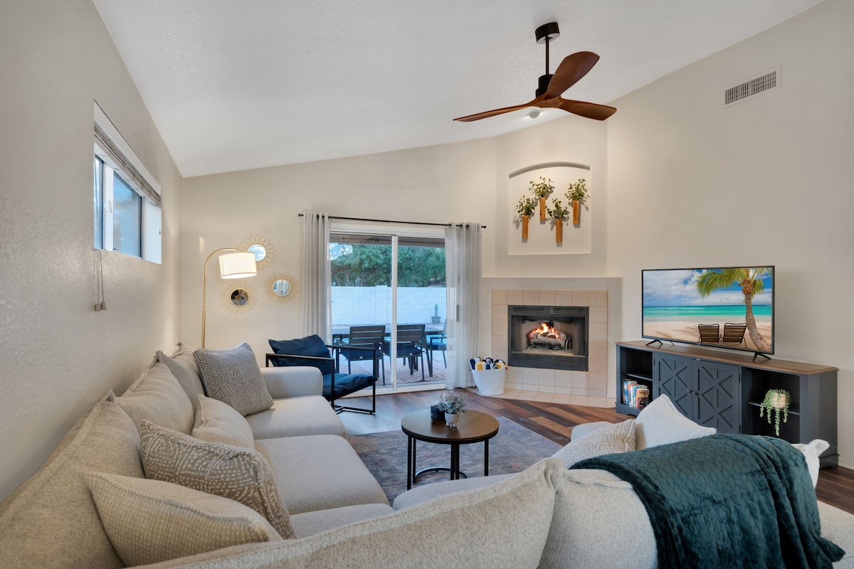 A cozy living area features a spacious sectional sofa and a circular table, creating an inviting space. A wall-mounted TV is positioned above a modern gas fireplace. Large windows allow natural light in, with sliding glass doors providing access to an outdoor seating area.