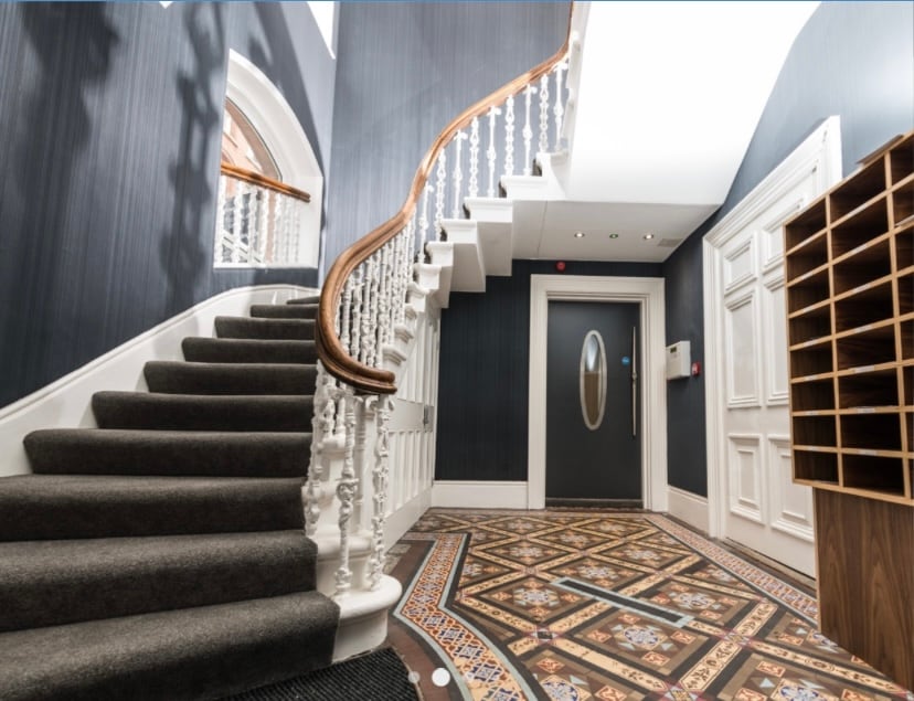 An elegantly designed staircase with a classic wooden handrail ascends to the upper level, complemented by intricately patterned tile flooring. The dark walls and polished features enhance the sophisticated ambiance of the welcoming entrance hall.