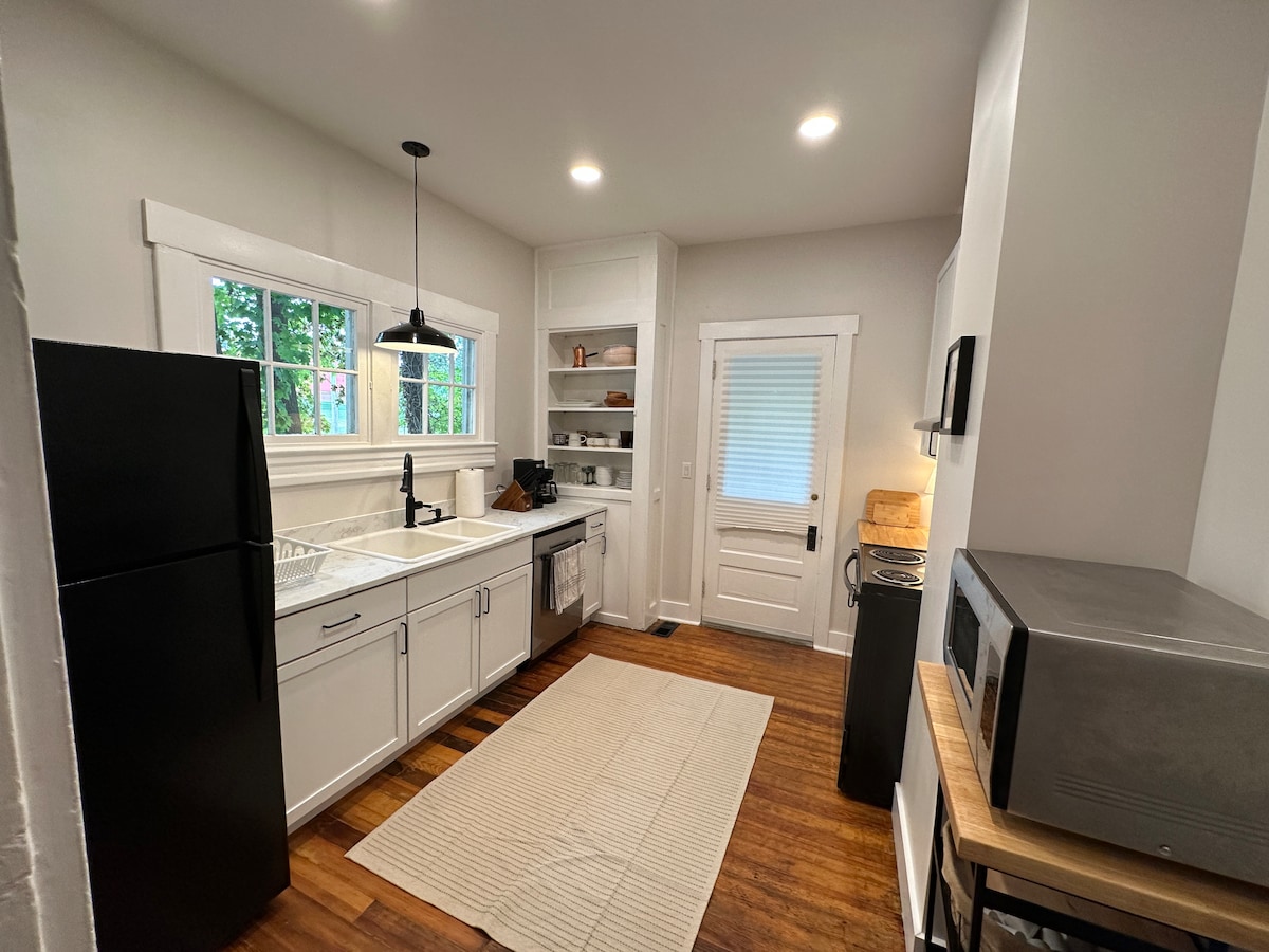 The kitchen features modern cabinetry and a spacious sink, accompanied by a black refrigerator and a microwave. Natural light enters through a row of windows, illuminating the wood flooring. A neutral area rug adds warmth to the space, enhancing its overall functionality.