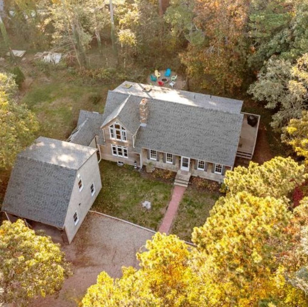 An aerial view of a spacious home surrounded by trees, showcasing a large gray roof and a charming pathway leading to the entrance. A detached garage is visible alongside, and colorful chairs are arranged on a deck, inviting relaxation outdoors.